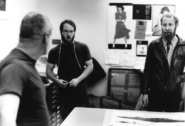 Artist Robert Stackhouse stands with artists Ray George and Richard Finch, reviewing prints laying on a table in front of them.